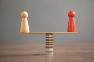 Uma foto em close-of de uma balança improvisada feita com uma régua de madeira equilibrada sobre uma pilha de moedas de metal. Em cada extremidade da régua, há um boneco de madeira estilo "pawn" (peão de jogo de tabuleiro): um de cor clara à esquerda e um vermelho à direita. O fundo é cinza neutro e a superfície é de madeira, transmitindo uma ideia de equilíbrio financeiro ou igualdade.