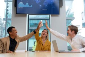 Três profissionais jovens — dois homens e uma mulher — sorriem e celebram sentados ao redor de uma mesa de escritório, unindo as mãos no alto em um gesto de "high five" triplo. Ao fundo, uma tela de TV exibe gráficos de negócios e um mapa mundial. O ambiente é moderno, com grandes janelas de vidro, sugerindo um momento de sucesso coletivo e trabalho em equipe.