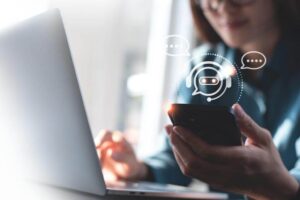 Close-up de uma mulher utilizando um notebook e um celular simultaneamente para realizar um atendimento via LiveChat. No centro da imagem, um ícone circular sobreposto ao smartphone exibe um atendente virtual com headset e balões de diálogo, ilustrando a integração do suporte híbrido e a jornada multicanal do usuário.