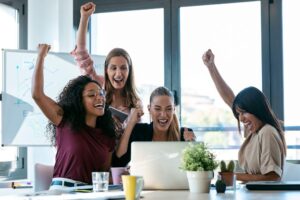 Quatro mulheres de uma equipe comercial comemoram juntas diante de um notebook em um escritório moderno, simbolizando o sucesso de uma cultura de vendas forte que gera receita previsível para o negócio.